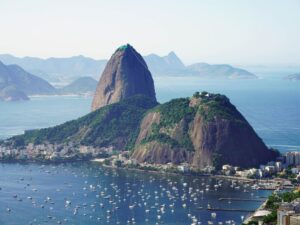 A panoramic view of Sugarloaf Mountain in Rio de Janeiro, with its iconic peak rising sharply from the Guanabara Bay. The ocean, dotted with numerous sailboats, reflects the sunny weather. Lush greenery covers the mountain's base while the cityscape nestles beneath a clear blue sky.