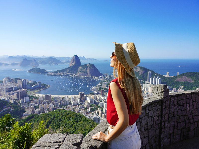 A woman in a straw hat and red top stands on a stone lookout, gazing over a panoramic view of Rio de Janeiro. The clear blue sky and calm waters create a serene backdrop, highlighting the vibrant urban and natural landscape nestled between mountains and ocean.