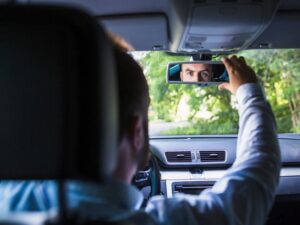 A man in a light blue shirt adjusts the rearview mirror of his rental car, focusing on his reflection. The car's interior is visible, showcasing a dashboard and air vents. Greenery outside suggests a lush, tree-lined road or park setting as he mentally runs through an inspection checklist.