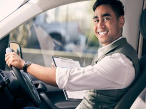 Man seated in a rental car, wearing a green vest over a white shirt. He is holding an inspection checklist clipboard and smiling at the camera. The interior of the car is visible, including the steering wheel. The blurred background outside suggests an outdoor setting.