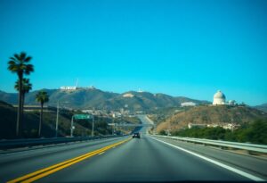 A clear Los Angeles highway scene with distant hills under a bright blue sky. The iconic Hollywood sign is visible on the left hillside. Surrounding the road are green bushes and palm trees, ideal for a road trip itinerary. A large white observatory dome crowns the right hilltop amid scattered buildings.