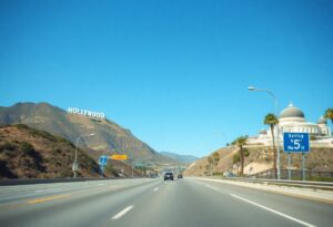 A highway stretches toward the iconic Hollywood sign on a hill under a clear blue sky, capturing the essence of a Los Angeles road trip. To the right is a large white observatory, while palm trees line the road. A green sign directs you to Interstate 5 amid sparse traffic with just a few cars visible.