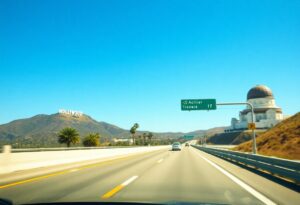 A clear view of a wide highway surrounded by dry hills under a blue sky sets the stage for an unforgettable Los Angeles road trip. The iconic Hollywood sign graces a hill to the left, while to the right, a dome-shaped observatory watches over. A green road sign marks the way ahead for your 3-day itinerary adventure.