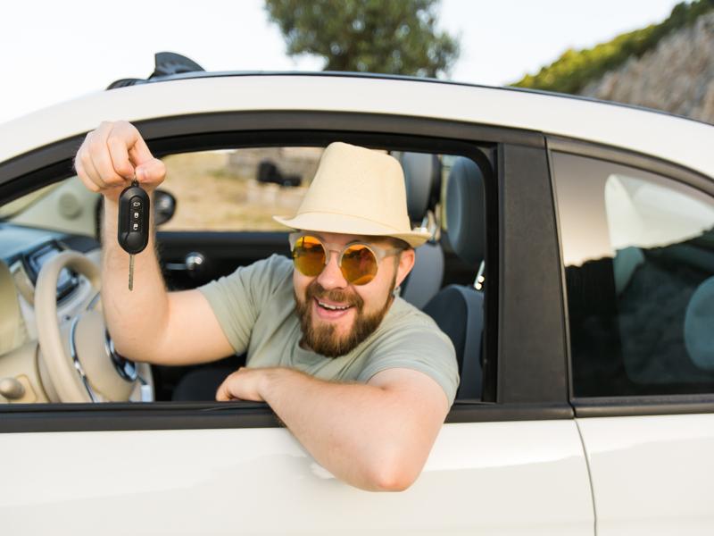 A man wearing a beige hat, sunglasses, and a light green shirt smiles from the driver's seat of a rental car. He holds a key fob out of the open window. The background shows a blurry landscape with trees and rocks, suggesting he has completed his checklist for the sunny adventure ahead.