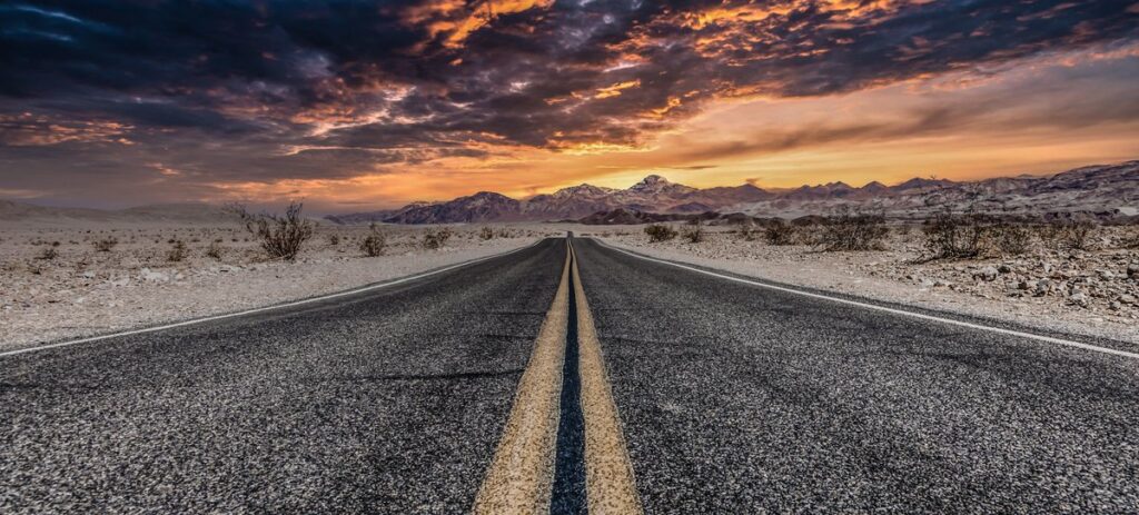 A straight road, reminiscent of Route 66, lined with a bright yellow line stretches towards distant mountains under a dramatic sky. The horizon features a vivid sunset with orange and purple clouds. Desert terrain with sparse shrubs flanks the road, enhancing the scene's vastness and rugged beauty.