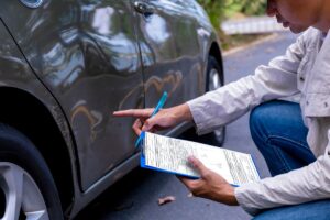 A person kneels on a road next to a gray car, pointing at a dent and scratch on the rear door. Holding a pen and clipboard, they might be noting details for an insurance claim or evaluating LDW coverage benefits. The parked car is in an outdoor setting surrounded by trees.