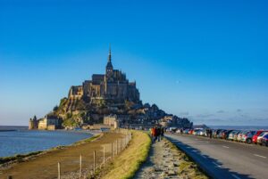 A distant view of Mont Saint-Michel against a clear blue sky showcases the medieval abbey and buildings atop a rocky island. As tourists walk along the path and parked cars line the route, road trip essentials become apparent for those driving in France to witness this stunning landmark surrounded by water.