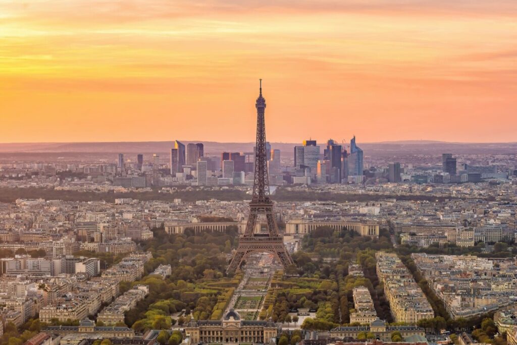 Aerial view of Paris at sunset, with the Eiffel Tower in the center. The skyline includes La Défense's skyscrapers, perfect for those planning a road trip across France. The sky glows with orange and pink hues, while historic buildings and green spaces dot the cityscape below.