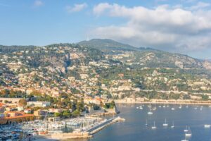 Scenic view of a coastal town with a marina filled with sailboats on a sunny day. Ideal for a road trip filled with driving in France, the town nestles against lush hills and rugged mountains under a partly cloudy sky. The coastline is dotted with villas, and a winding road traces the shore.