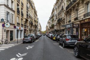 A narrow urban street bustling with parked cars lines both sides, showcasing the charm of European architecture with balconies. Shop signs, like "Roy Chocolats," stand out under an overcast sky. Bicycle lanes are clearly marked for safe cycling—a nod to the unique rules of driving in France.