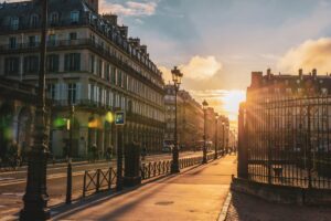 As the sun sets over a European city, its golden light dances across classical buildings, casting long shadows. Bicyclists and pedestrians navigate the streets with ease, reminiscent of a road trip in France. Decorative street lamps and ornate iron gates complete the picturesque scene under a partly cloudy sky.