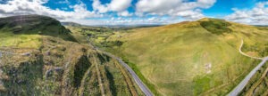 A panoramic view of a green, hilly landscape under a partly cloudy blue sky unfolds. A winding road, essential for those driving in Ireland, cuts through the scene. Rolling hills with patches of grass and scattered bushes define the foreground, while distant hills and a valley extend to the horizon.