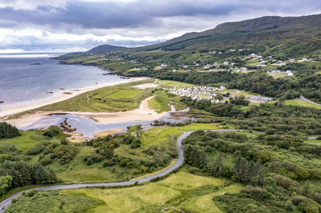 Aerial view of a coastal landscape in Ireland, with a winding road perfect for driving adventures. Lush greenery and a sandy beach with a small lagoon line the way. Houses are scattered along the hillside, mountains in the backdrop, as the sea extends under a cloudy sky.