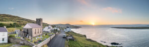 Enjoy a panoramic view of a coastal village at sunset, perfect for those curious about driving in Ireland. A narrow road winds through stone buildings, including a church with a tower. Grassy hills rise on the left while the sea sparkles on the right, as the sun casts its warm glow over this picturesque scene.