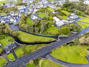 Aerial view of a small residential area featuring neatly arranged houses surrounded by lush green lawns and trees. A winding road, akin to those found when driving in Ireland, cuts through the scene, bordered by driveways and a small stream with a footbridge. The landscape is a mix of open grassy areas and clustered homes.