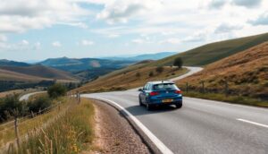 A blue car from an unlimited mileage car rental drives on a winding road through a scenic hilly landscape. Grassy hills fill the foreground, while rolling green mountains and a partly cloudy sky grace the background. A fence lines the left side, creating a peaceful and picturesque atmosphere.