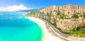 Scenic view of a coastal town with buildings perched on a cliff overlooking a turquoise sea. A winding road, perfect for an Italian road trip, runs along the base of the cliff beside a white sandy beach. Green hills are visible in the background under a clear blue sky.