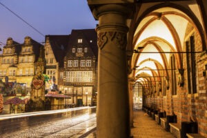 View of a cobblestone street with historic buildings at dusk, reminiscent of a scene where tips for driving in Germany might come in handy. The brick and stone structures boast intricate details, large windows, and decorative facades. An arched walkway with lamps stretches alongside, as a blurred tram leaves light trails on the wet street.