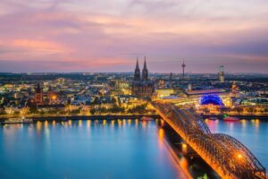 Aerial view of Cologne, Germany, at dusk reveals the illuminated Hohenzollern Bridge spanning the Rhine River, guiding drivers mindful of local rules towards the majestic Cologne Cathedral with its twin spires. City lights twinkle on the water as pink and orange hues paint a picturesque urban landscape.