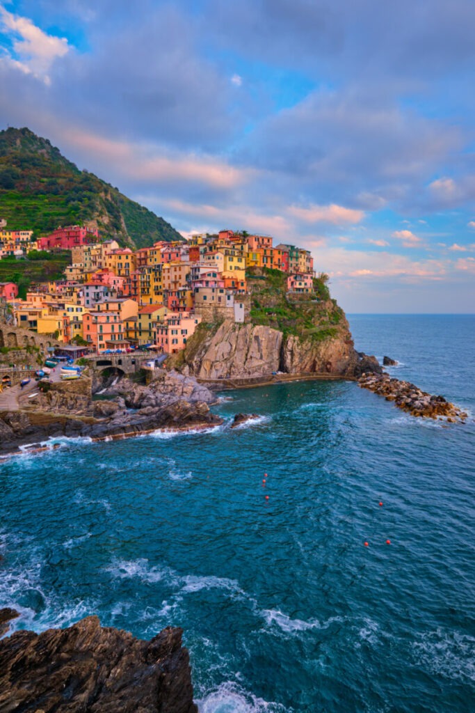 Colorful houses perched on a cliff overlook the blue sea in Manarola, Cinque Terre—a must-see on any Italian road trip. The vibrant buildings are surrounded by lush green hills under a partly cloudy sky. The rocky shoreline breaks the waves, adding texture to the picturesque coastal scene.