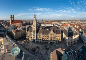 Aerial view of Munich's Marienplatz, featuring the Gothic-style New Town Hall with its tall spire. The Frauenkirche, with its distinctive twin domes, is visible in the background. As you navigate these bustling streets, keep a Germany driving guide handy for a seamless journey under the clear blue sky.