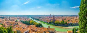 Panoramic view of Verona, Italy, perfect for an Italian road trip. The Adige River gracefully curves through the city, showcasing a skyline of historic buildings and prominent towers under a clear blue sky. Lush green trees along the riverbank contrast with terracotta rooftops.