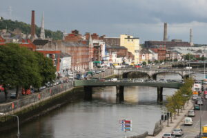 A bustling urban scene with a river on the left, bordered by a stone embankment and lined with trees. A bridge crosses the river, connecting streets filled with cars—a must-know for driving in Ireland—and colorful buildings. In the distance, tall chimneys and spires rise against a cloudy sky.