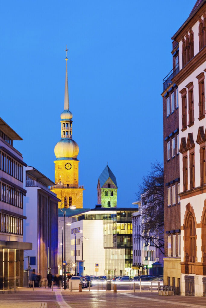 Street view at dusk presents a historic church with a tall, illuminated spire and clock tower. Modern buildings flank the church, contrasting old and new architecture. As you navigate these streets while driving in Germany, the deep blue sky and warm lights add a serene ambiance.