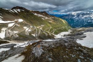 A winding mountain road cuts through a rugged landscape in the Alps. Snow patches dot the rocky terrain and green slopes. Dark clouds loom above, with distant peaks visible under a hint of blue sky, creating a dramatic and scenic view perfect for an Italian road trip.