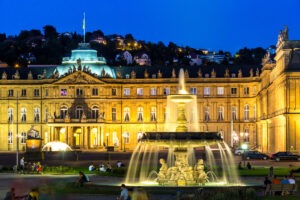 An illuminated baroque palace glows warmly at night, its intricate facade highlighted by soft lights. A grand fountain with statues and cascading water takes center stage, surrounded by people. Hills dotted with lit houses rise in the background, much like the orderly charm of driving in Germany.