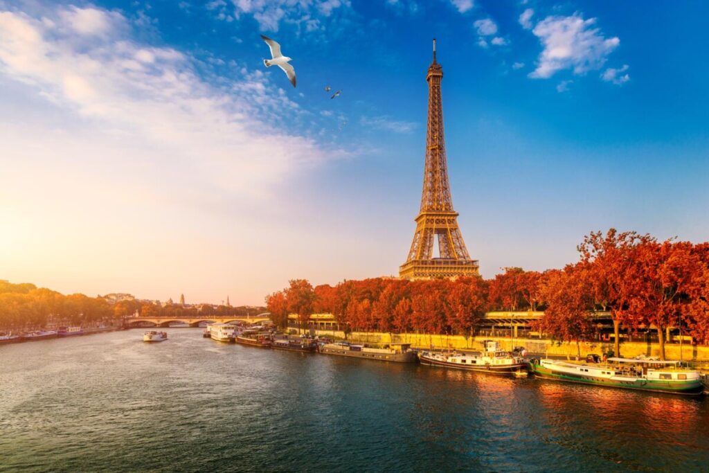 The Eiffel Tower at sunset with a clear blue sky and scattered clouds paints the perfect European holiday destination. The Seine River reflects golden light, boats moored along its banks. Vibrant autumn trees line the riverbank as a seagull gracefully flies overhead, adding motion to the scene.