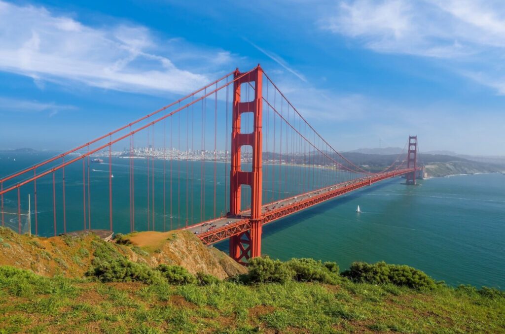 Experience the ultimate road trip as you take in a scenic view of San Francisco's iconic Golden Gate Bridge under a clear blue sky. The striking red suspension bridge stretches across the water with sailboats below, framed by green shrubs and a grassy hill, while the city's skyline dots the horizon.