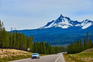 Embark on the ultimate road trip as your car glides along a paved road flanked by lush green forests. In the distance, a snow-capped mountain towers under the blue sky, offering a glimpse into nature's boundless beauty—a true journey from San Francisco to Seattle.