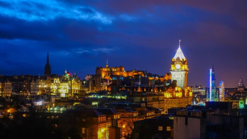 Night view of Edinburgh, showcasing its skyline with illuminated historic buildings. A prominent clock tower shines brightly against a deep blue sky. In the background, Edinburgh Castle is lit up, creating a striking silhouette with attractions aplenty as city lights reflect on the streets below.