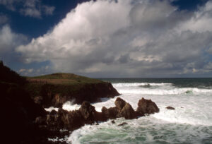 The rocky coastline unfurls like a hidden gem along a winding road trip from Seattle to San Francisco, where waves crash against the cliffs beneath a partly cloudy sky. Grassy hilltops rise against the turbulent ocean, and dark rocks dominate the choppy seascape stretching toward the distant horizon.