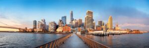 Panoramic view of Seattle’s skyline at sunset with modern skyscrapers and a prominent suspension bridge on the left. The buildings reflect golden light, while a long wooden pier leads from the foreground, guiding the eye toward this iconic urban landscape—a perfect road trip destination.