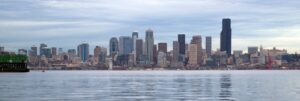 Panoramic view of a city skyline across calm water, featuring numerous tall buildings with varying architectural designs. The sky is overcast with light gray clouds, creating a serene atmosphere. This ultimate experience from San Francisco to Seattle promises breathtaking sights along the way.