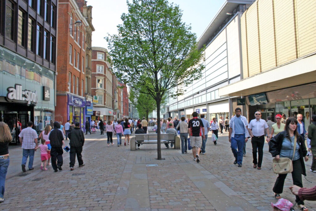 A bustling urban street scene with pedestrians walking on a cobblestone pathway, perfect for Every Traveler. Buildings line both sides, featuring shops with signs like "allsaints." A leafy tree offers shade over a bench. People are dressed casually, enjoying one of the unmissable experiences on a sunny day.