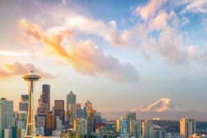 Seattle's skyline at sunset, highlighted by the Space Needle and Mount Rainier in the background, offers a serene scene perfect for any road trip experience. City buildings glow with warm sunlight as pink and orange clouds dot the sky, creating a picturesque view.